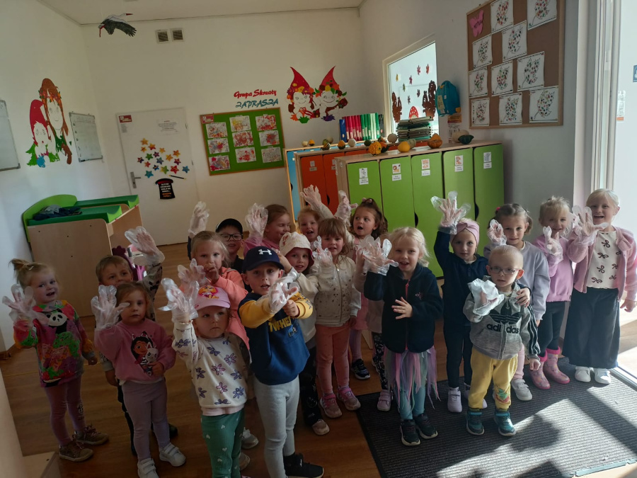 Grupa przedszkolaków stoi w sali przy kolorowych szafkach. Dzieci mają na dłoniach jednorazowe rękawiczki i radośnie pokazują je w stronę aparatu. W tle widoczne są jesienne dekoracje