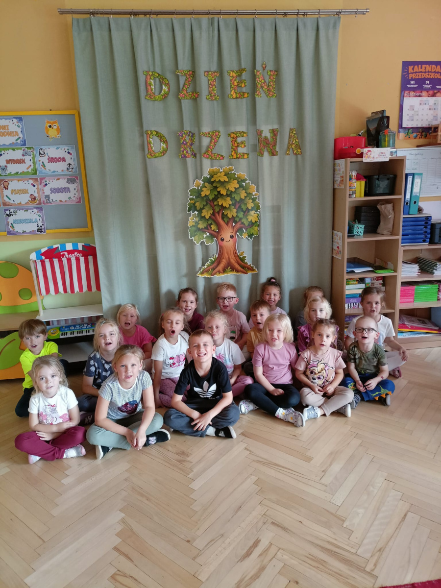 Grupa dzieci siedzi na podłodze w sali przedszkolnej przed dekoracją z napisem Dzień Drzewa i ilustracją uśmiechniętego drzewa pozując do wspólnego zdjęcia
