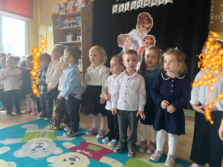 Dzieci w eleganckich strojach stoją w szeregu przed dekoracją z okazji Dnia Edukacji Narodowej prezentując występ artystyczny