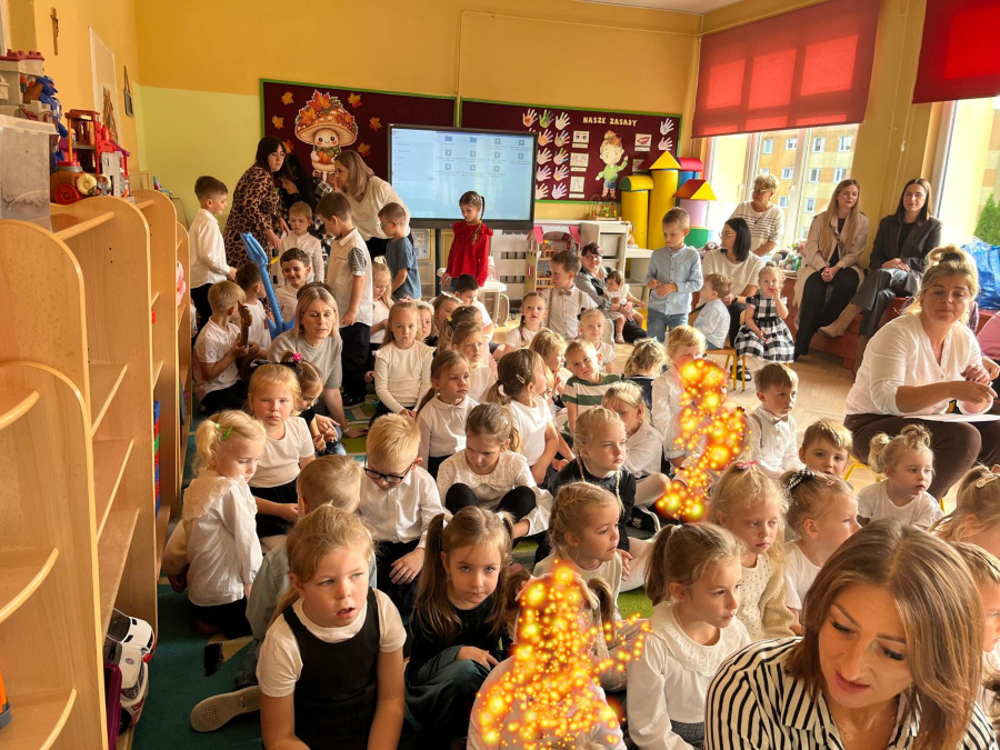 Dzieci w odświętnych strojach siedzą na dywanie podczas uroczystości z okazji Dnia Edukacji Narodowej. W sali obecne są również kobiety
