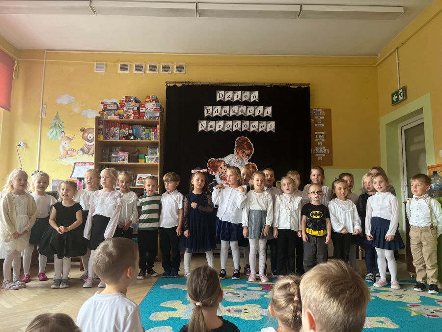 Dzieci w odświętnych strojach stoją przed dekoracją z napisem Dzień Edukacji Narodowej i śpiewają podczas uroczystości przedszkolnej
