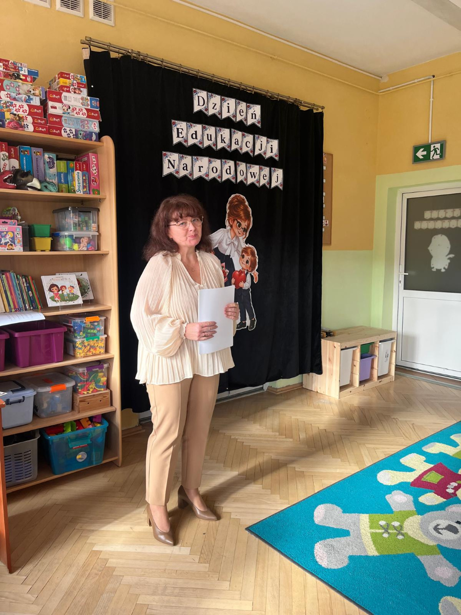 Kobieta w jasnej bluzce i beżowych spodniach stoi przed dekoracją z napisem Dzień Edukacji Narodowej trzymając w dłoniach kartki