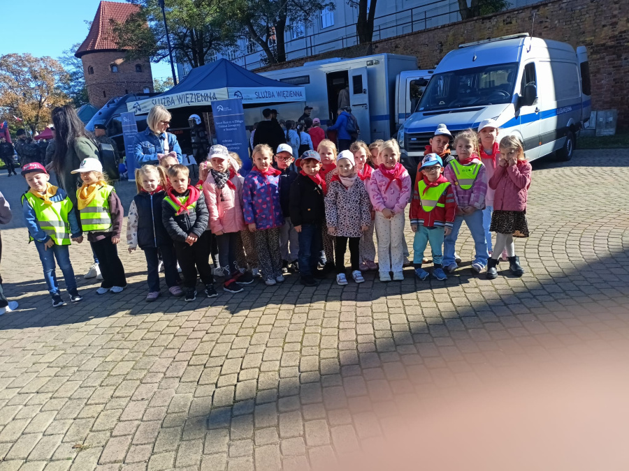 Grupa przedszkolaków pozuje do wspólnego zdjęcia podczas wydarzenia plenerowego na tle pojazdów Służby Więziennej i stoiska informacyjnego