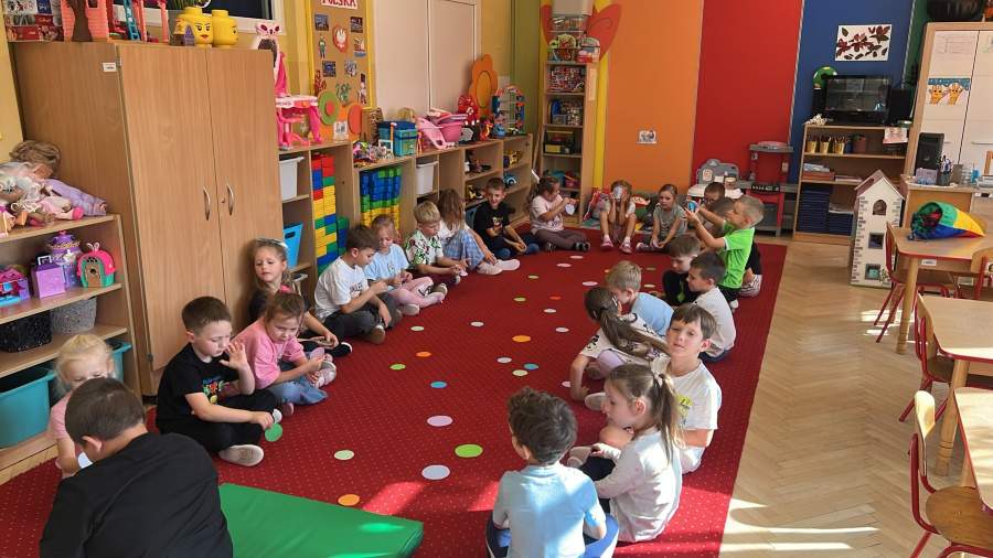 Dzieci siedzą w dwóch rzędach na dywanie, pomiędzy kolorowymi kołami ułożonymi na podłodze, uczestnicząc we wspólnej zabawie ruchowej.