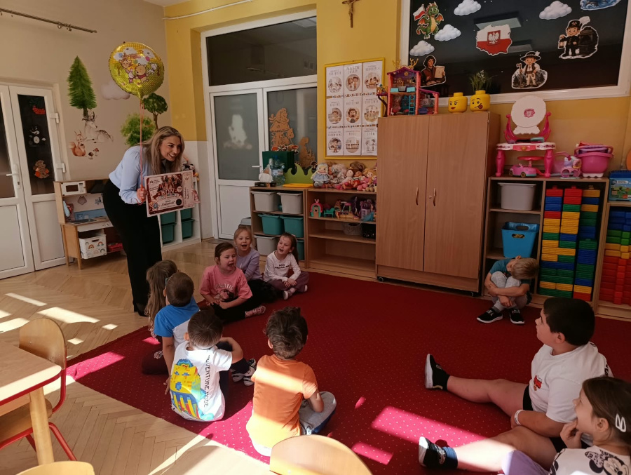 Dzieci siedzą na dywanie i uczestniczą w zajęciach prowadzonych przez kobietę która prezentuje im książkę oraz kolorowy balon