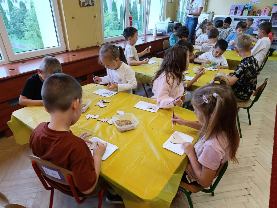 Dzieci siedzą przy stolikach i wykonują prace plastyczne podczas warsztatów ozdabiając drewniane elementy związane z tematyką bursztynu