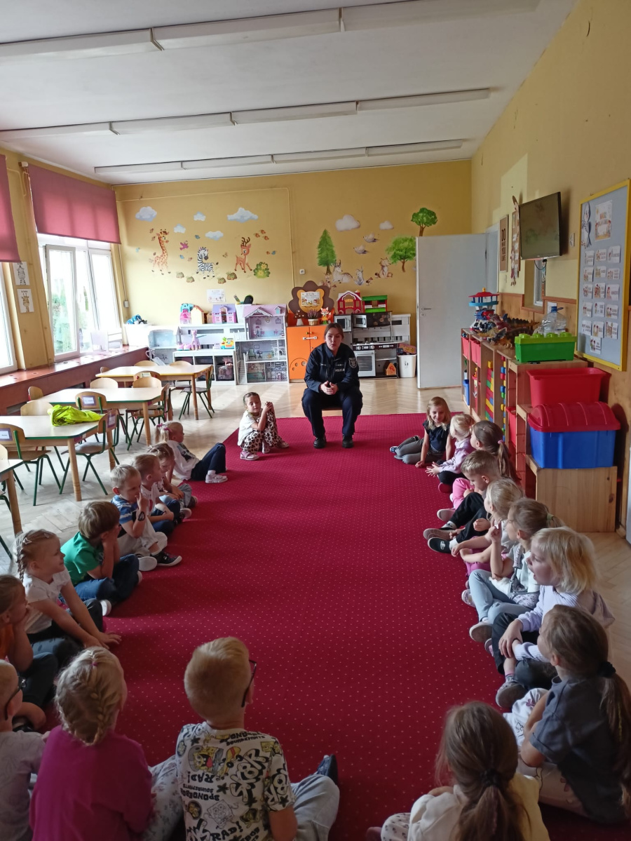Dzieci siedzą na dywanie w sali przedszkolnej i uczestniczą w spotkaniu z policjantką poświęconym zasadom bezpieczeństwajpeg