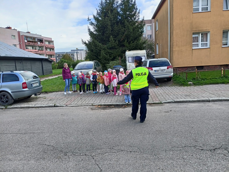 Policjantka w kamizelce odblaskowej pokazuje grupie przedszkolaków, jak prawidłowo i bezpiecznie przechodzić przez ulicę.