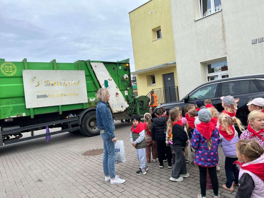 Grupa dzieci obserwuje śmieciarkę podczas zajęć edukacyjnych na terenie przedszkola, poznając pracę służb komunalnych i zasady dbania o środowisko